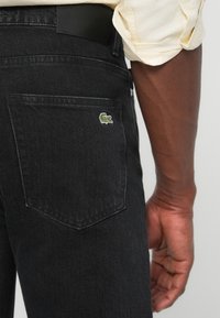 Bolsillo trasero de pantalones vaqueros negros con el logotipo del cocodrilo de Lacoste, usados por una persona de piel oscura y camisa de manga enrollada color crema.