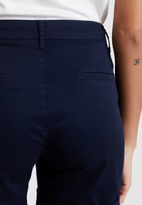 Laivastonsiniset housut, joissa on sileä pinta, keskitason vyötärö, takataskut ja hillityt ompelukoristeet.