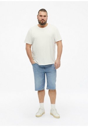 Man met baard en naar achter gebonden haar, die een wit T-shirt, blauwe denimshorts, witte sokken en witte sneakers draagt, staand met handen in de zakken.