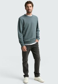 Grijsblauwe crewneck trui van een gestructureerde stof, gedragen over een wit overhemd, gecombineerd met donkere jeans en witte sneakers. Staande pose op een lichte achtergrond.