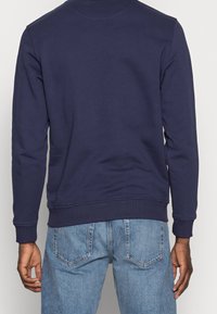 Sudadera azul marino con cuello redondo, puños y dobladillo acanalados, con textura suave. Combinada con vaqueros azul claro, mostrando vistas posteriores.