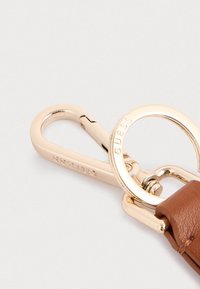 Porte-clés en cuir marron avec un clip carabiner en métal doré et un anneau circulaire, comportant une gravure de la marque sur le matériel.