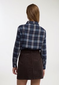 Camicia di flanella a quadri blu e grigia con maniche lunghe e collo, abbinata a una gonna di velluto a coste marrone scuro. Dettagli di trama e motivo evidenti.