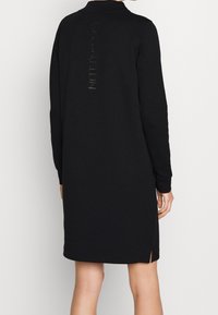 Černé sweatshirtové šaty s dlouhými rukávy, límečkem, bočními rozparky a lesklým logem na zadní straně. Měkká, strukturovaná tkanina.