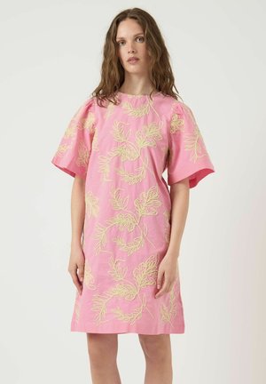 Kvinde med langt, bølget hår iført en løs lyserød kjole med gul blomstret broderi og korte pufærmer mod en ensartet lys baggrund.