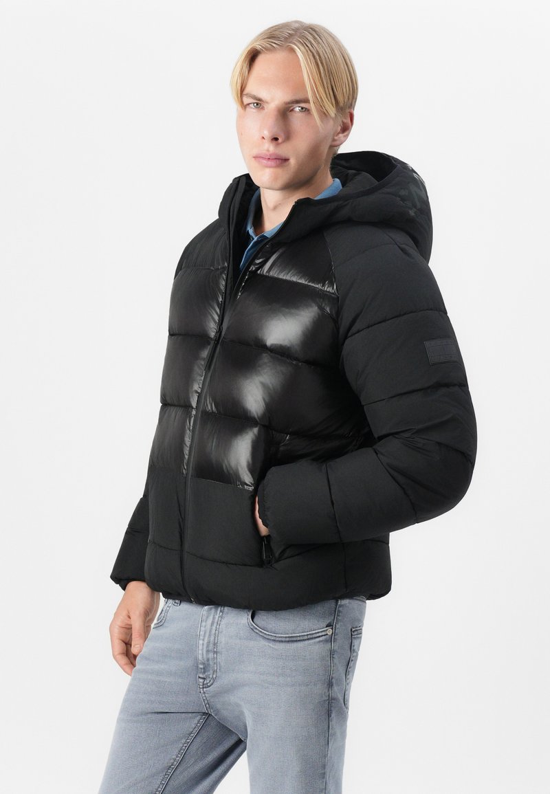 Schwarze Daunenjacke mit glänzender Oberfläche, Reißverschluss vorne, Kapuze und gerippten Bündchen. Kombiniert mit hellgrauen Jeans.