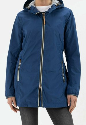 Personne portant une veste à capuche bleue avec fermetures éclair à l'avant et sur les côtés, associée à un pantalon gris foncé, debout devant un fond uni.