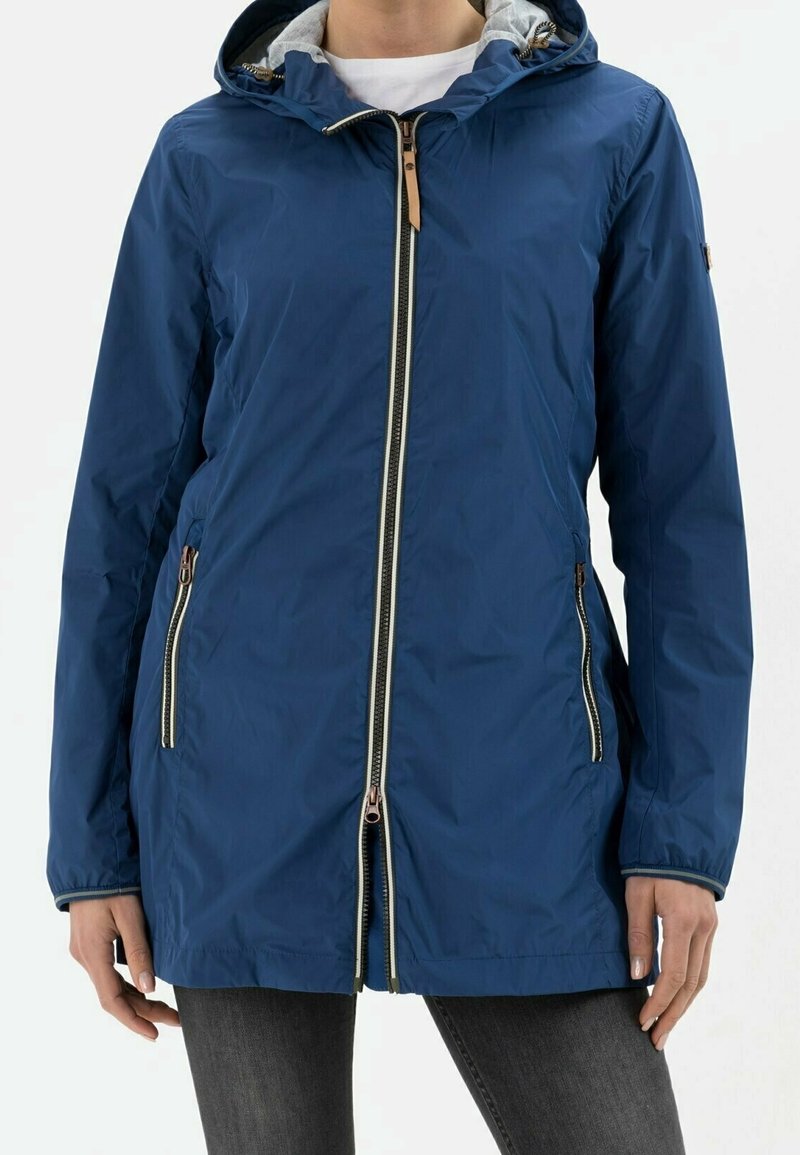 Personne portant une veste à capuche bleue avec fermetures éclair à l'avant et sur les côtés, associée à un pantalon gris foncé, debout devant un fond uni.