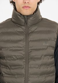 Vaahto vest tumman oliivinvihreänä korkealla kauluksella, etuvetoketjulla ja huomaamattomalla brändäyksellä rinnassa. Sileä, kiiltävä pinta ja vaakasaumat.