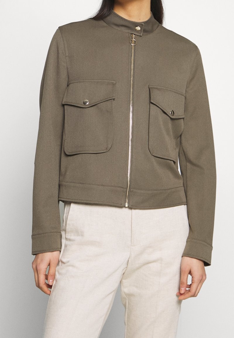 Femme portant une veste zippée vert olive avec deux poches à rabat et un pantalon beige clair sur un fond uni.