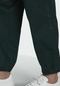 Gambe inferiori che indossano pantaloni jogger neri con polsini elasticizzati e sneakers bianche su uno sfondo grigio chiaro.