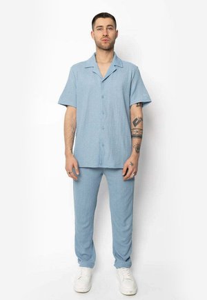 Chemise à manches courtes bleu clair avec col et boutons à l'avant, associée à un pantalon décontracté assorti, en tissu texturé. Baskets blanches.