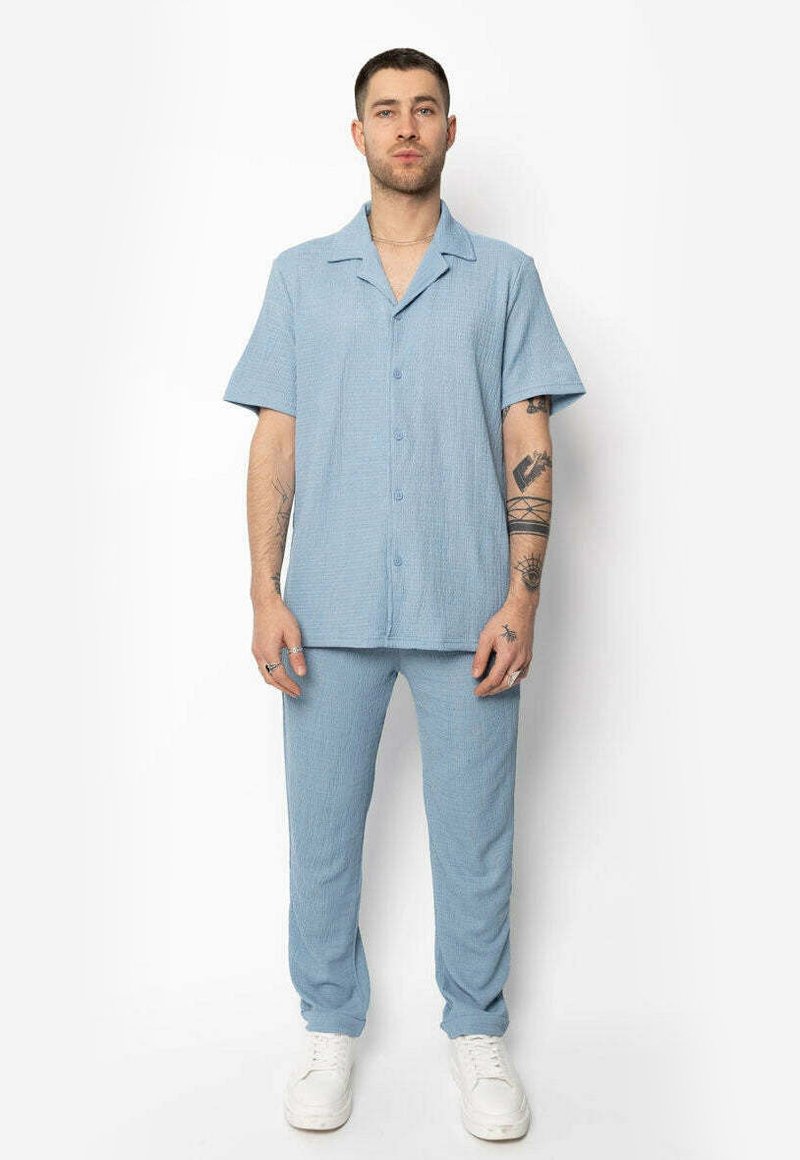 Chemise à manches courtes bleu clair avec col et boutons à l'avant, associée à un pantalon décontracté assorti, en tissu texturé. Baskets blanches.