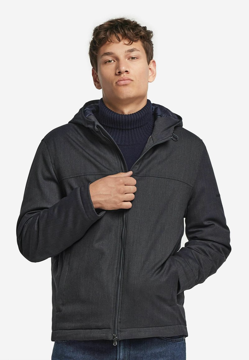 Ung man som bär en mörkgrå huva-jacka och marinblå polotröja, med en hand som justerar jackans dragkedja, står mot en vit bakgrund.