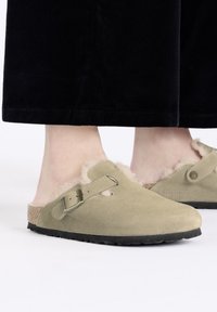 Zuecos de ante verde claro con un forro interior de felpa, tira trasera y detalle de hebilla. Suela de goma negra y resistente. Diseño de perfil bajo.