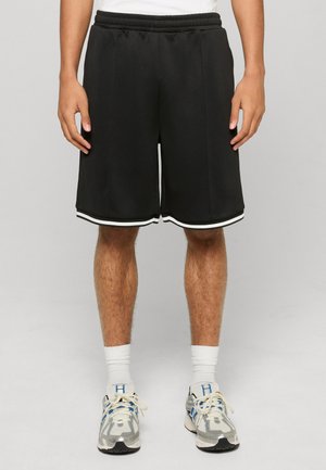 Mann trägt schwarze athletische Shorts bis zum Knie mit weißem Besatz, weiße Socken und graue und blaue Laufschuhe, steht vor einem schlichten Hintergrund.
