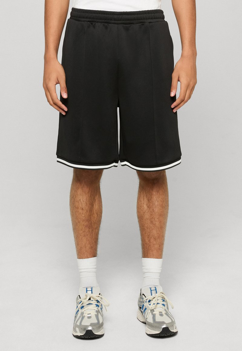 Homme portant un short de sport noir longueur genou avec un liseré blanc, des chaussettes blanches et des chaussures de course grises et bleues, debout devant un fond uni.