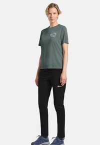 T-shirt vert à manches courtes avec un petit design graphique sur la poitrine gauche, associé à un pantalon noir et des chaussures de sport bleues.
