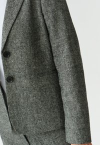 Šedý blazer vyrobený z texturované tkaniny, s dvěma černými knoflíky a jednou náprsní kapsou, s mírně přiléhavým střihem.