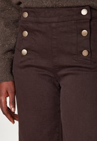 Pantaloni marroni a vita alta con quattro bottoni metallici sul davanti e un tessuto liscio e texturizzato. Presentano tasche laterali.