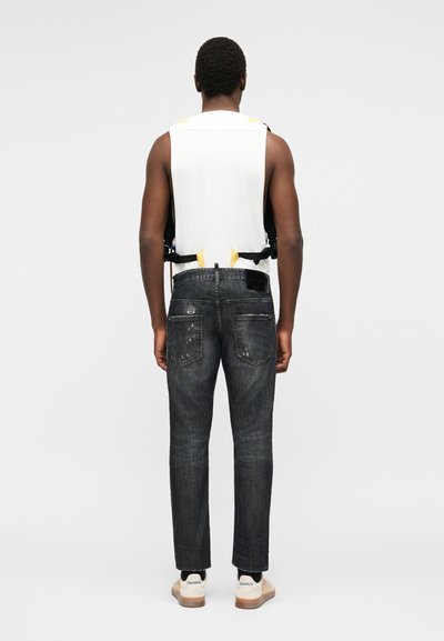 Άνδρας με την πλάτη γυρισμένη φοράει λευκό φανελάκι με μαύρους ιμάντες, μαύρα φθαρμένα τζιν και λευκά αθλητικά παπούτσια σε απλό ανοιχτό φόντο.