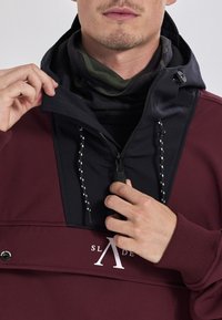 Bordeaux hoodie med en sort lynlåspanel, justerbare snore og et logo. Indersiden har et mørkt mønstret tørklæde.