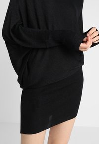 Svart stickad tröja med långa ärmar och ribbade mudd, tillsammans med en figurnära svart ribbad minikjol. Båda plaggen har en slät yta.