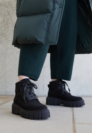Bottes noires épaisses à plateforme portées avec un pantalon court vert foncé et une doudoune noire sur un sol carrelé.