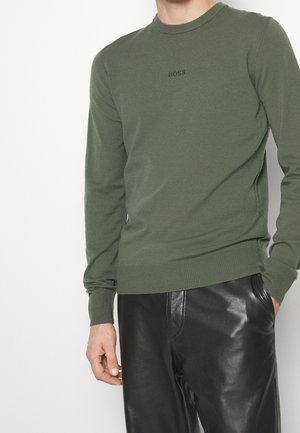 Mand iført olivengrøn sweater med teksten "BOSS" og sorte læderbukser, står med den ene hånd i lommen mod en ensfarvet baggrund.