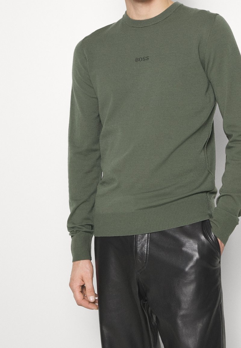 Homme portant un pull vert olive avec le texte "BOSS" et un pantalon en cuir noir, debout avec une main dans la poche devant un fond uni.