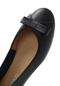 Black leather ballet flat featuring a rounded toe, a small bow accent on the vamp, and a beige interior lining. Smooth texture throughout.