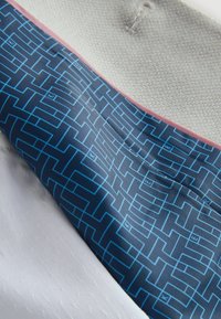 Oggetto tessile con tessuto bianco testurizzato e una fascia a motivo blu con linee geometriche, bordata da un sottile rifinitura rosa.