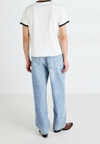 Persona con una camiseta blanca con ribetes negros y jeans ligeros de corte holgado, de pie de espaldas a la cámara sobre un fondo blanco liso.