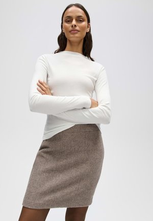 Vrouw met lang donker haar die een wit topje met lange mouwen en een bruine geruite rok tot op de knieën draagt, staat met gekruiste armen en kijkt zelfverzekerd.