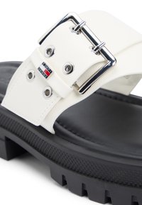 Πέδιλο με χοντρή μαύρη σόλα, χοντρό λευκό δερμάτινο λουρί, ασημένια αγκράφα και ετικέτα με το λογότυπο Tommy Jeans κοντά στις τρυπητές οπές.