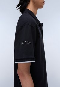 Polo shirt nera a maniche corte, caratterizzata da un accento bianco a contrasto sui polsini e un logo sulla manica sinistra. Tessuto dalla texture liscia.