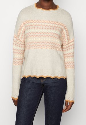 Personne portant un pull crème avec des motifs géométriques pêche, des bords festonnés orange, associé à un jean bleu foncé sur un fond uni.