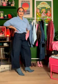 Femeie cu machiaj albastru, pulover bleu deschis și pantaloni închiși la culoare, pozând lângă un suport de haine, în fața unui perete verde cu postere colorate.