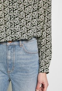 Blouse à manches longues avec un motif géométrique noir et blanc sur un fond vert menthe, accompagnée de jeans en denim bleu clair.