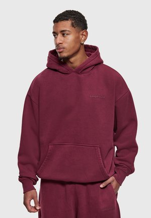 Noormees, kes kannab sobivaid burgundiapunaseid kapuutsiga pusa ja teksapükse, seisab ühe käega taskus ja vaatab küljele tavalise tausta ees.