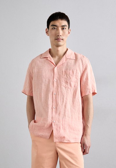 Chemise à manches courtes, boutonnée, en lin corail clair avec une poche poitrine et une coupe décontractée. Le mannequin porte un pantalon de couleur pêche. Texture lisse.