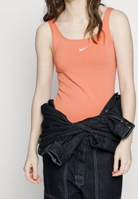 Orange figurerad linne med fyrkantig halsringning och tunna band, prytt med en vit Nike-logotyp. Mörk denimjacka knuten runt midjan.