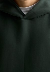 Felpa verde scuro con una texture morbida e liscia; presenta una grande tasca frontale e un cappuccio con cordino. Sotto, è visibile una maglietta bianca a girocollo.