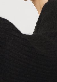 Svart stickad tröja med ett texturerat mönster och vida ärmar, delvis visad på en axel mot en neutral bakgrund.