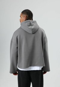 Grijze hoodie gemaakt van zachte stof, met een grote capuchon, brede mouwen en een cropped ontwerp. Draagt over een wit shirt met zwarte broek.