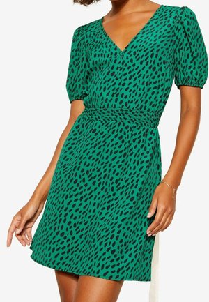 Mujer con vestido corto verde con manchas negras irregulares, mangas cortas abullonadas, escote en V y cintura fruncida.