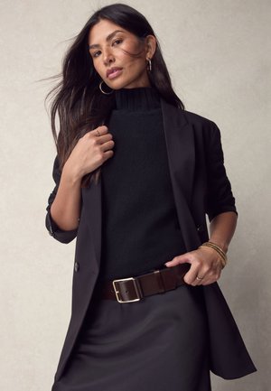 Mujer con cabello largo y oscuro que lleva un suéter de cuello alto negro, blazer y falda a juego, cinturón marrón, pendientes de aro dorados y pulseras.
