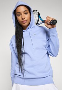 Hoodie albastru deschis din amestec de bumbac, cu o buzunare frontală, glugă cu cordon și un logo verde pe piept. Modelul ține o rachetă de tenis.