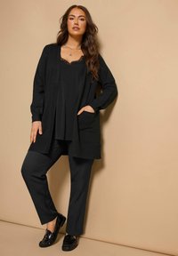 Schwarze Strickjacke mit offenem Vorderteil und Taschen, über einem schwarzen Oberteil mit Spitzenbesatz getragen, kombiniert mit schwarzen Hosen und glänzenden schwarzen Slippern.