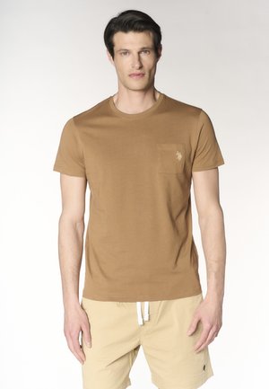 Uomo che indossa una maglietta marrone a maniche corte con tasca e pantaloncini beige con coulisse, in piedi davanti a uno sfondo semplice.